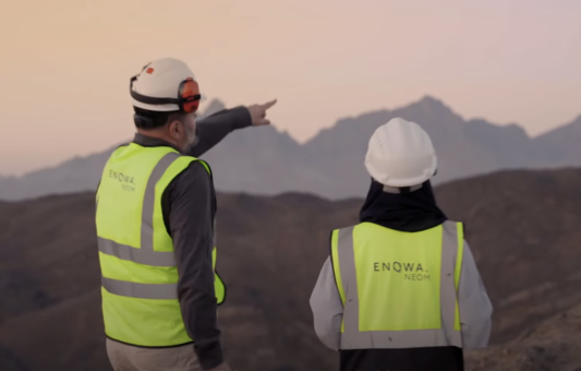 Two people wearing safety helmets and ENOWA NEOM high-visibility vests stand outdoors at dusk; one points towards distant mountains, symbolizing how ENOWA is revolutionizing construction in Saudi Arabia.